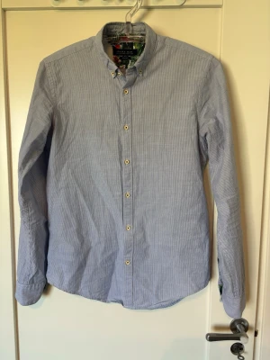 Blå- och vitrandig skjorta från Zara - Klassisk blå- och vitrandig skjorta från Zara med button-down krage och vita knappar. Långärmad modell i bomull med snyggt mönstrat tyg på insidan av manschetterna. Perfekt för en stilren look. Storlek är M men sitter som en S