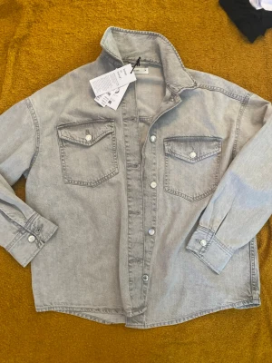 Grå jeansjacka  - Snygg grå jeansjacka från Gina tricot med klassisk krage, två bröstfickor med lock och knappar samt knäppning framtill. Jackan har raka ärmar med manschett och en avslappnad passform. Perfekt för en cool och avslappnad stil.
