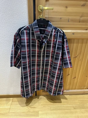 Rutig kortärmad skjorta i bomull - Snygg kortärmad skjorta med rött, svart, vitt och grått rutmönster. Klassisk krage och knäppning framtill, samt bröstficka. 