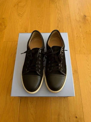  Lanvin  - Svarta skor från Lanvin med ovandel i mocka och tåparti i slätt läder. Skorna har svarta skosnören, metallöljetter och vit sula med Lanvin-logga undertill. Snygg och stilren design som funkar till många olika outfits. Storlek 44 