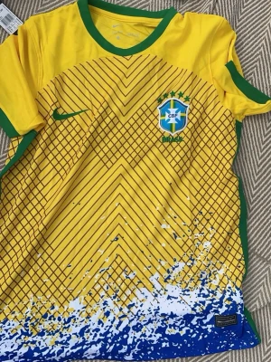 Brasilien matchtröja Nike gul - Säljer en gul Brasilien matchtröja från Nike med gröna detaljer vid krage och ärmslut. Tröjan har ett grafiskt mönster i svart.