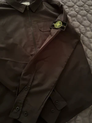 Svart overshirt från Stone Island - Säljer en svart overshirt från Stone Island med klassisk krage, bröstficka och dragkedja framtill. Ikonisk logopatch på vänster ärm. Materialet är slätt och lätt, perfekt för lager-på-lager. Snygga knappar vid ärmslut och stilren design.