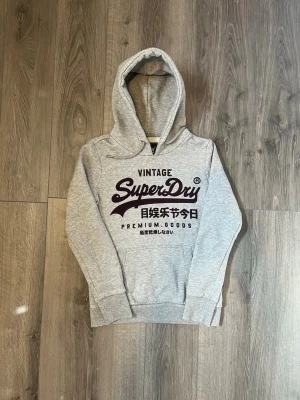Grå Superdry hoodie med tryck - Snygg grå hoodie från Superdry med stor vinröd logga och japanskt tryck på bröstet🤗 Passar snyggt för de som har Xs🙌🏻 säljer då tröjan var för liten för mig, har alltså inte använt den alls.