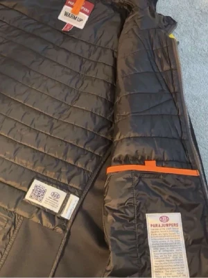 Svart parajumper cardigan  - Snygg svart pufferjacka från Parajumpers med huva och dragkedja framtill. Jackan har quiltad design, orange detaljer och klassisk Parajumpers-logga på ärmen. Tillverkad i lätt och slitstarkt material som håller värmen under kalla dagar.