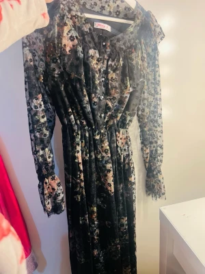 Blommig långklänning från Holly - Supersnygg långklänning från Holly med blommigt mönster i mörka toner. Klänningen har långa ärmar, knappar framtill, volangdetaljer vid axlarna och ett bälte i midjan. Perfekt för dig som vill sticka ut med en romantisk och trendig look.