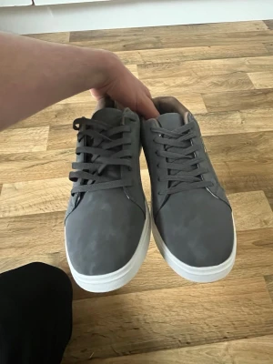 Gråa sneakers med vit sula - Snygga grå sneakers med klassisk låg profil och vit sula. Skorna har matchande grå skosnören och en stilren design utan synliga loggor. Materialet ser ut att vara slätt syntet eller mockaimitation, vilket ger en modern look.