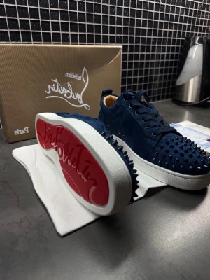 Blåa Louboutin sneakers med nitar - Snygga mörkblå sneakers från Christian Louboutin i mocka med ikoniska röda sulor och silverfärgade nitar över hela ovandelen. Skorna har vita sulor och klassisk låg profil. Perfekta för dig som vill sticka ut med en edgy och lyxig stil.