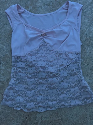 Ljusrosa spetsbody med rosett - Söt ljusrosa body med breda axelband och dekorativ rosett framtill. Nedre delen är i genomskinlig blommig spets som ger en romantisk känsla. Överdelen är i mjukt tyg och har en feminin v-ringning. 