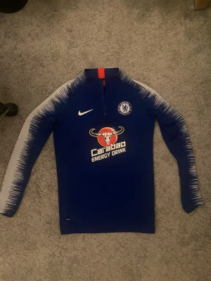 Chelsea träningströja från Nike - Blå långärmad träningströja från Nike med Chelsea FC-logga på bröstet och Carabao Energy Drink-tryck på framsidan. Jackan har halv dragkedja och vita detaljer längs ärmarna med coolt mönster. Perfekt för fotbollsträning eller sport.