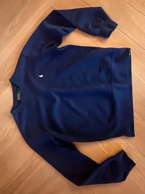 Mörkblå sweatshirt från Polo Ralph Lauren - Klassisk mörkblå sweatshirt från Polo Ralph Lauren med rund halsringning och den ikoniska vita logon broderad på bröstet. Tröjan har ribbade muddar vid ärmslut och nederkant, perfekt för en avslappnad och stilren look. 