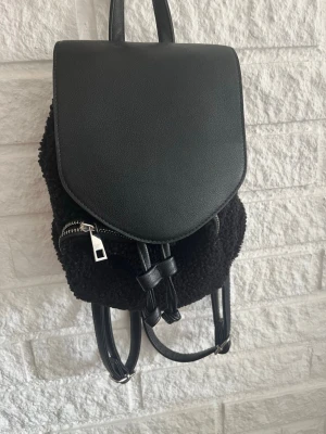 Svart ryggsäck med teddy och läderdetaljer - Snygg svart ryggsäck med lock i fejkläder och kropp i mjukt teddy-material. Justerbara axelband i svart läderimitation och dragkedja framtill. Perfekt för dig som vill ha en trendig och praktisk väska med unik textur.