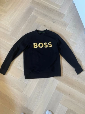 Svart sweatshirt från BOSS - Svart sweatshirt från BOSS med stor gul logga på bröstet. Tröjan har rund halsringning och långa ärmar. Tillverkad i mjukt material som känns skönt mot huden. Perfekt för en avslappnad och stilren look.