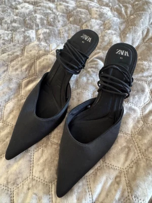 Svarta spetsiga sandaler från Zara - Säljer ett par svarta sandaler från Zara med spetsig tå och flera tunna remmar över foten. De har en stilren design och är tillverkade i ett tygmaterial som ger en snygg och modern look. Perfekta för dig som gillar trendiga skor med attityd.