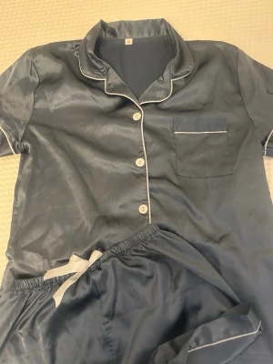 Blå satinpyjamas med vita detaljer - Snygg blå pyjamas i glansigt satinmaterial med kortärmad skjorta och matchande shorts. Skjortan har vita knappar, vit passpoal längs krage och ficka samt en bröstficka. Shortsen har resår i midjan och vit dragsko. Perfekt för chillkvällar hemma.