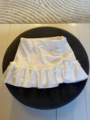 Vit kortkjol med volang och knyt - Supersöt vit kortkjol med volangkant och knyt i sidan. Kjolen har en lätt struktur i tyget och ger en fräsch och somrig vibe. Från gina i storlek S.