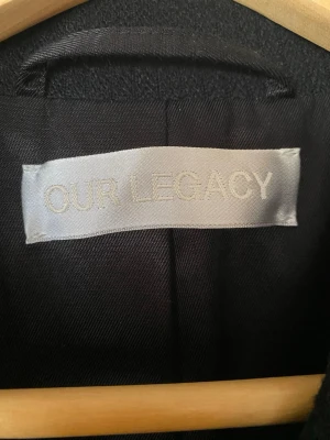 Svart ullkappa från Our Legacy - Stilren svart ullkappa från Our Legacy med klassisk krage och knäppning framtill. Kappan har två snedställda fickor och en rak, lång siluett som ger en tidlös look. Perfekt för dig som gillar minimalistisk och modern design.