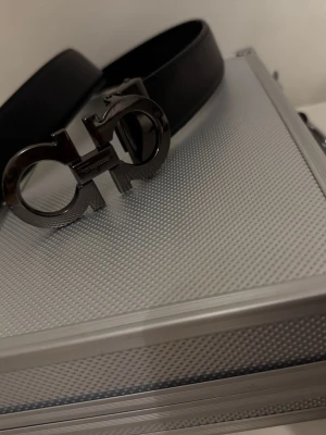 Svart silver skärp från Salvatore Ferragamo - Snyggt svart silver skärp från Salvatore Ferragamo med ikoniskt dubbel-Gancini-spänne i mörk metall. Bältet är gjort i slätt skinn och har en stilren, modern look som passar till många outfits.