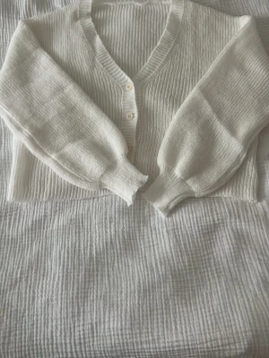 Vit ribbad kofta  - Säljer en vit ribbstickad kofta med v-ringning och ballongärmar. Koftan har tre små knappar framtill och breda muddar vid ärmsluten. Perfekt att slänga över en topp för en chill look. Den är aldrig använd och är från h&m. Tröjan är liten i storlek. 