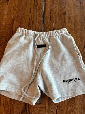 Essentials Fear of God shorts grå - Säljer ett par ljusgrå shorts från Essentials Fear of God med snörning i midjan och logotyp tryckt på benet. De har en relaxed passform och är tillverkade i mjuk bomull, perfekta för en chill och avslappnad stil. Stl XS men passar S