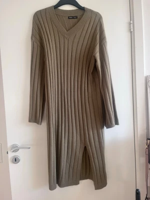 Beige/grön ribbad klänning  - En beige/grön klänning med ribbad struktur och v-ringad hals. Klänningen har långa ärmar och en slits framtill som ger en snygg detalj. Perfekt för dig som gillar en enkel och stilren look.