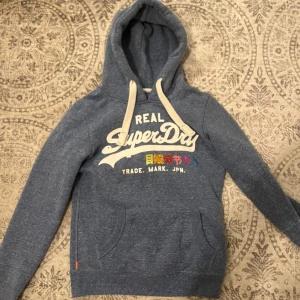 Superdry blå hoodie - STORLEK: S.  SKICK: 9/10.                                       Säljer denna sällsynta superdry hoodien med flerfärgade tecken på grund av att den är för liten för mig men den är i välldigt fint skick.