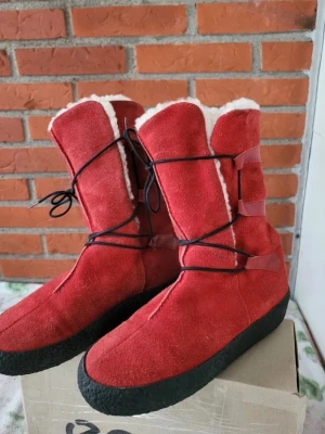 Röda mockaboots från Catwalk Stockholm 37 - Säljer ett par röda moon boots med svart snörning och grov sula. Bootsens utsida är i mocka och insidan är fodrad med mjuk, vit syntetpäls. Perfekta för kalla vinterdagar och ger en cool retrovibe.