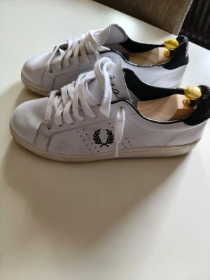 Vita Fred Perry sneakers med svart detalj - Snygga vita sneakers från Fred Perry med svart häl och klassisk lagerkrans-logga på sidan. Skorna har vita snören, perforerade detaljer och är tillverkade i läder. Perfekta för en stilren och sportig look.