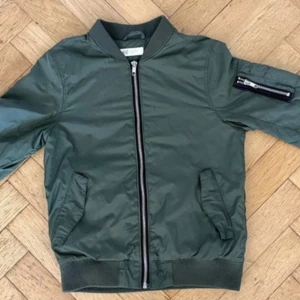 Grön bomberjacka från H&M - Snygg militärgrön bomberjacka från H&M med klassisk ribbad krage, muddar och dragkedja framtill. Jackan har fickor på sidorna och en extra ficka med dragkedja på ärmen. Yttermaterialet är i polyester och insidan är mjuk och skön. Finns två mindre revor som på baksidan av höger ärm som inte påverkar funktionen och inget som syns tydligt.