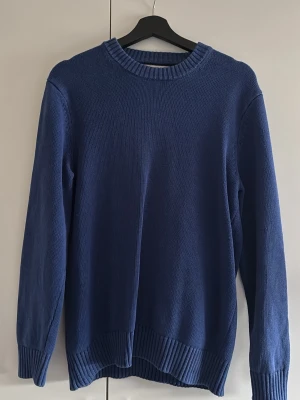 Blå stickad tröja med rund hals - En enkel och stilren blå stickad tröja med rund halsringning och ribbade muddar vid ärmslut och nederkant. Perfekt för lager på lager och passar till det mesta i garderoben. Mjuk och bekväm, i klassisk modell med lång ärm.
