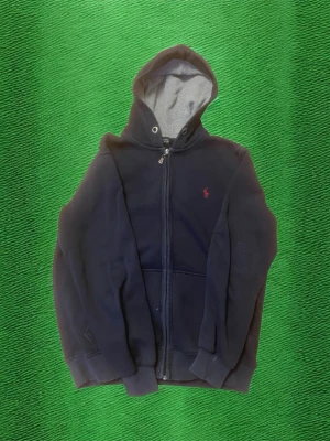 Mörkblå hoodie från Polo Ralph Lauren - Säljer en mörkblå hoodie med dragkedja från Polo Ralph Lauren. Den har en huva med grått innerfoder, ribbade muddar och fickor på framsidan. Klassisk röd Polo-logga broderad på bröstet. Perfekt för en chill och stilren look. Passar bra till small