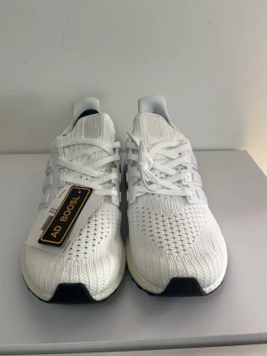 Vita Adidas Ultraboost sneakers - Fräscha vita Adidas Ultraboost sneakers med stickad ovandel och klassiska tre ränder på sidan. Skorna har en bekväm Boost-sula och vita skosnören. Perfekta för dig som gillar stilrena och sportiga sneakers.