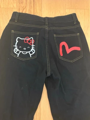 Svarta Hello Kitty jeans - Unika svarta jeans med kontrastsömmar och coola tryck på bakfickorna – Hello Kitty på ena och ett rött hjärta på den andra. Rak passform och klassisk midja. Perfekta för dig som vill sticka ut med en lekfull och trendig stil. Kortmodell passar mig precis som är 163 cm. Aldrig använda!❤️