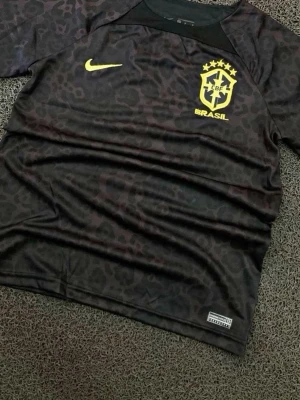 Svart Brasilien fotbollströja Nike - Snygg svart fotbollströja från Nike med Brasilien-emblem och gul Nike-logga på bröstet. Tröjan har ett diskret leopardmönster och är tillverkad i ett lätt, andningsbart material som passar perfekt till träning eller match.