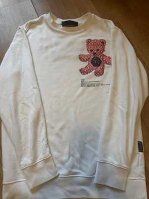 Vit sweatshirt med nalle från Plein - Vit sweatshirt från Philipp Plein med en rosa nalleapplikation på bröstet och textdetaljer. På ryggen står det 'PLEIN Teddy' i rosa brodyr tillsammans med en liten döskalle. Tröjan har rund halsringning och ribbade muddar. Perfekt för dig som gillar streetwear med en twist.
