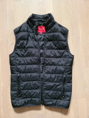 Svart dunväst från Guess XS - Snygg svart dunväst från Guess med quiltad design och hög krage. Västen har dragkedja framtill, två fickor med dragkedja och diskret Guess-logga på bröstet. Perfekt för lager-på-lager och streetstyle.