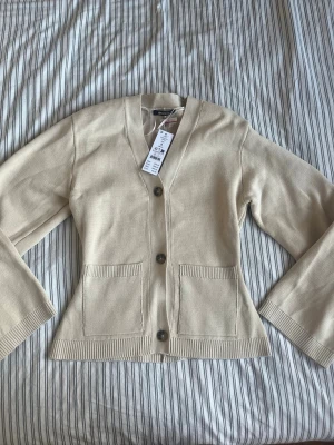 Gina tricot - Stilren beige kofta från Gina tricot med v-ringning och tre knappar framtill. Koftan har två fickor fram och är tillverkad i mjukt stickat material. Perfekt för lager-på-lager och enkel att matcha med olika outfits.
