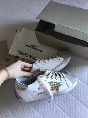 Golden Goose vita sneakers med glitterstjärna - Golden Goose sneakers i vitt läder med en glittrig guldstjärna på sidan och silverfärgade detaljer vid hälen. Klassisk låg modell med vita snören och grå mockadetaljer på tån. Snygg och trendig design som sticker ut. Storlek 40 men känns mer som 39.