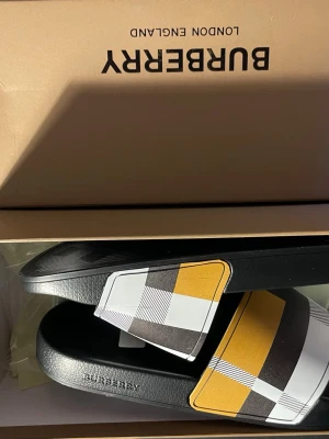 Tofflor med rutigt mönster - Snygga tofflor  med klassiskt rutigt mönster i svart, vitt, brunt och gult. Tofflorna har en bred rem över foten och svart sula i syntetmaterial. Perfekta för en trendig och avslappnad stil.