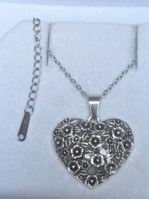Halsband med hjärthänge i silverfärg - Snyggt handgjort halsband i silverfärgad metall med ett stort hjärthänge dekorerat med detaljerade blommor. Hänget lyser lila i mörker efter att ha fått dagsljus eller solljus på sig. Kedjan är tunn och justerbar med förlängningskedja i rostfritt stål. Hänget har en vintagekänsla och är perfekt för dig som gillar unika smycken med romantisk touch.