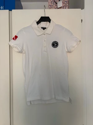 Vit pikétröja från GANT med emblem - Vit pikétröja från GANT med klassisk krage, knappar framtill och broderat emblem på bröstet. Röd detalj på ena ärmen. Tillverkad i mjuk bomull och har korta ärmar. Perfekt för en clean och sportig look.