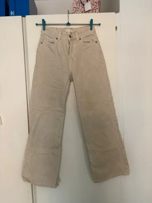 Beige raka jeans med hög midja - Säljer ett par beige jeans med raka ben och hög midja. Klassisk femficksmodell med bredare passform i benen. Jeansen har bälteshällor och stängs med knappar. Perfekta för en avslappnad och trendig look.