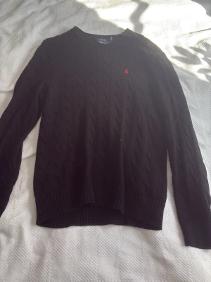 Svart stickad tröja Polo Ralph Lauren - Svart stickad tröja från Polo Ralph Lauren med klassiskt kabelstickat mönster och rund hals. Tröjan är äkta med nypris på cirka 2000kr. Liten defekt på ryggen men syns inte jättetydligt. Skriv för fler funderingar!👍
