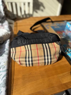 Burberry shoppingväska med klassiskt mönster - Säljer en snygg shoppingväska med det ikoniska beige rutmönstret och svarta detaljer. Väskan har ett brett axelband, dragkedja. Det finns någon liten fläck men det syns inte tydligt. Matchande keps finns också! 