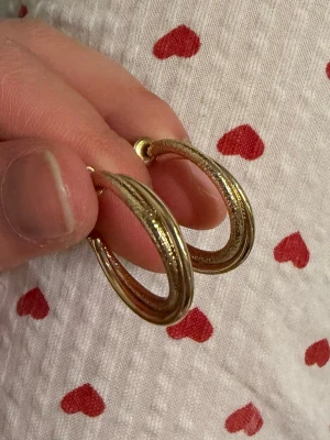 Guldiga creoler med graverat mönster - 100% NICKELFRIA ENLIGT MÄRKET, NYPRIS 79kr. Har inte kvar lapp men aldrig använda. Snygga örhängen i form av creoler i guldfärgad metall. De har ett graverat mönster längs hela ytan som ger en lyxig och trendig känsla. Perfekta för att lyfta vilken outfit som helst med lite extra bling. Stift och pluppar i matchande färg.