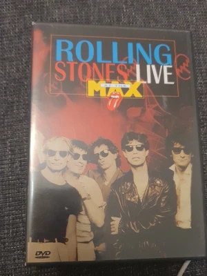 Rolling Stones Live DVD - DVD med Rolling Stones livekonsert 'At The Max'. Omslaget har bandets logga och färgerna svart, röd, blå och gul. Innehåller klassiska låtar som 'Paint It Black' och 'Satisfaction'. Perfekt för dig som gillar rock och samlar på musikfilmer.