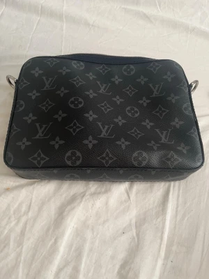 Louis Vuitton svart monogram handväska - Snygg svart handväska från Louis Vuitton med klassiskt monogrammönster i grått. Väskan har en rektangulär form och är tillverkad i tåligt läder. Perfekt accessoar för dig som vill ha en exklusiv och trendig look. Dustbag och originalkartong medföljer.  Axelremen saknas.