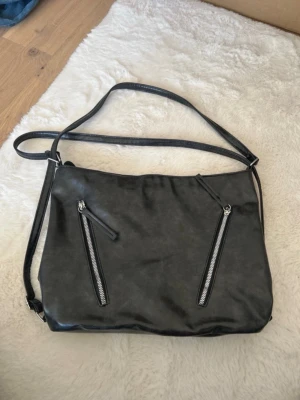 Svart axelväska med dragkedjor - Snygg svart axelväska i konstläder med två sneda dragkedjor framtill och justerbar axelrem. Väskan har ett stort huvudfack med ljusrosa tygfoder och flera innerfickor för småsaker. Perfekt för dig som vill ha en stilren och praktisk väska.