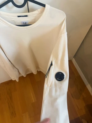 Vit sweatshirt från C.P. Company - Vit sweatshirt från C.P. Company med rund halsringning och lång ärm. Tröjan har den ikoniska linsen på ärmen samt en liten ficka med dragkedja. Tillverkad i mjukt bomullsmaterial för en clean och stilren look.