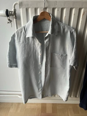 A Days March - Säljer en ljusblå skjorta i linne med korta ärmar och klassisk krage. Skjortan har knappar framtill och en bröstficka. Perfekt för en avslappnad och stilren look.