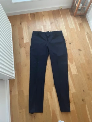 Marinblåa kostymbyxor med smal passform - Snygga marinblåa kostymbyxor med smal passform och klassisk design. Byxorna har hällor för bälte, två fickor fram och två bakfickor med knapp. Perfekta för en stilren look. Materialet ser ut att vara slätt och dressat. Slim fit